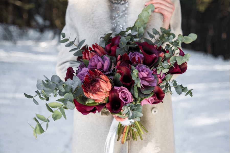bride in snow holding qinter birdal bouquet with colors of deep purple and red - Forever Bridal Wedding Shows