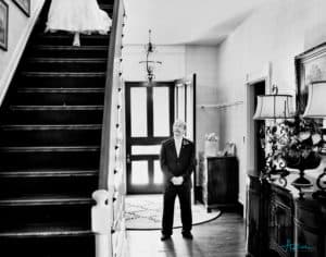 Bride walks down the stairs as her father patiently waits to see her in the wedding dress for the first time.