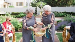Two older women in dresses hold a basket of flowers at a wedding.