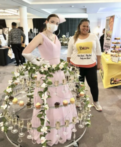 woman wearing wedding dress with cupcakes placed around them. Another woman stands behind her and smiles